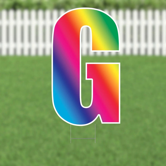 Colorful yard sign letter H with a rainbow gradient fill and white outline, displayed against a white picket fence and green lawn background.
