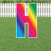 Colorful yard sign shaped like the letter I with a vibrant rainbow gradient displaying pink, orange, yellow, green, cyan, and blue colors, displayed on grass with a white picket fence background.