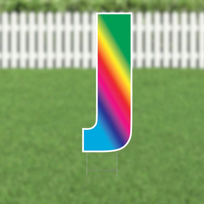 Colorful yard sign letter J with a rainbow gradient transitioning from green at the top through yellow, orange, red, pink, magenta, and blue at the bottom, displayed on grass with a white picket fence background.