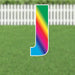 Colorful yard sign letter J with a rainbow gradient transitioning from green at the top through yellow, orange, red, pink, magenta, and blue at the bottom, displayed on grass with a white picket fence background.