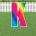 Large yard sign letter K with vibrant rainbow gradient from blue to pink to green, displayed on grass with white picket fence background.