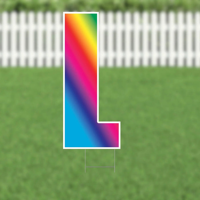 Colorful yard card letter M with a vibrant rainbow gradient transitioning from red and yellow at the top through blue and cyan at the bottom, displayed on green grass with a white picket fence background.