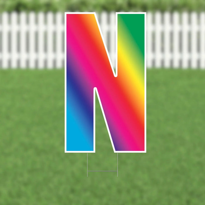 Colorful yard card letter N with rainbow gradient design featuring pink, orange, yellow, green, and blue sections, displayed on green grass with white picket fence background.