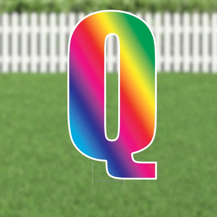 Oversized letter Q yard sign with a vibrant rainbow gradient fill and white outline, displayed against a green lawn and white picket fence background.