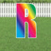Colorful yard sign letter R with a rainbow gradient fill and white outline, displayed against a white picket fence and green grass background.