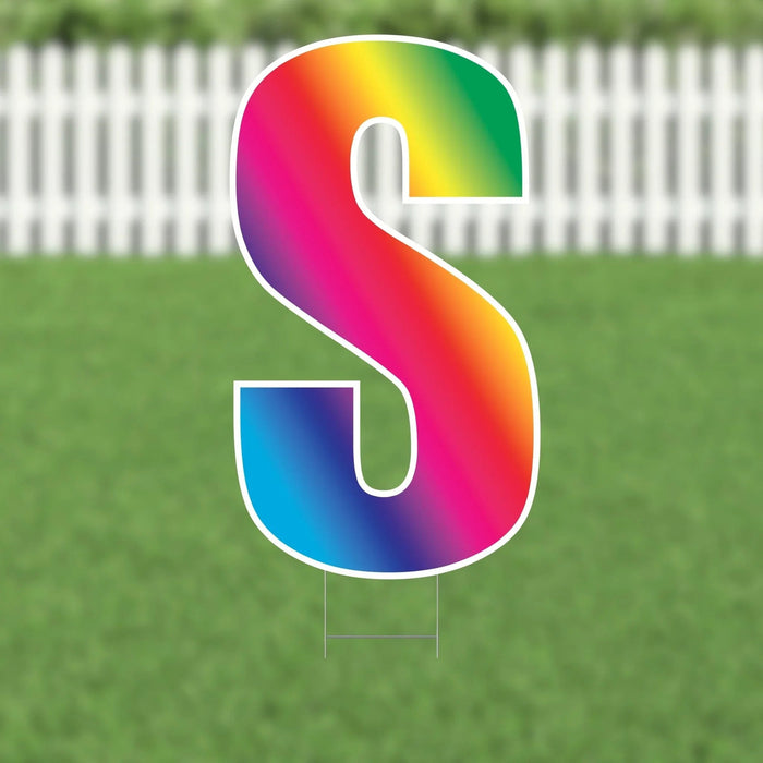 Rainbow gradient letter S yard sign with white outline displayed on green grass against white picket fence background.