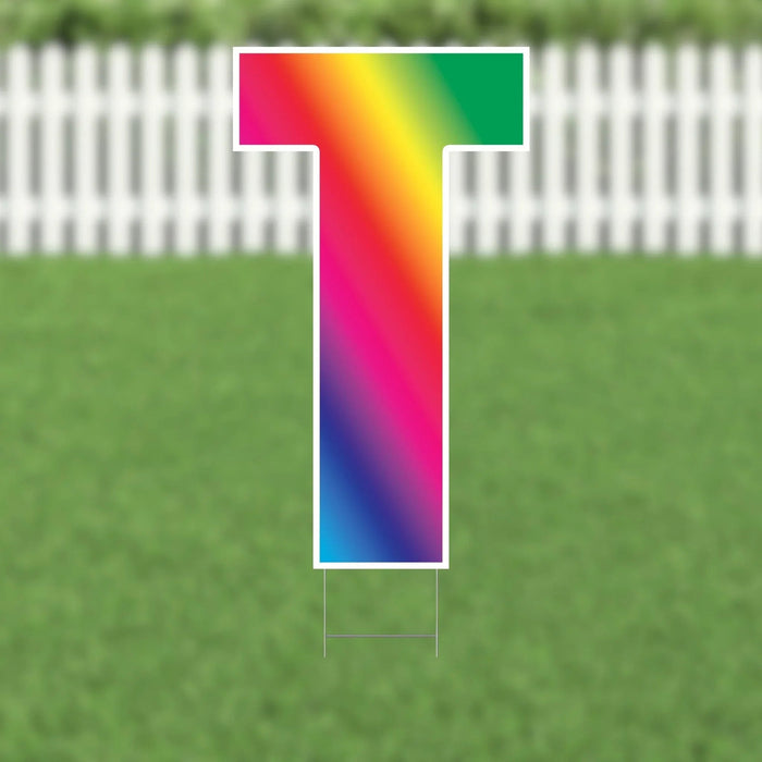 Colorful yard sign letter T with a rainbow gradient transitioning from pink and red at the top through yellow and green to blue and purple at the bottom, displayed against a white picket fence and green grass background.