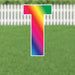 Colorful yard sign letter T with a rainbow gradient transitioning from pink and red at the top through yellow and green to blue and purple at the bottom, displayed against a white picket fence and green grass background.