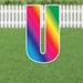Colorful letter V yard sign with a rainbow gradient transitioning from pink and red at the top through yellow, green, and blue to purple at the bottom, displayed on a lawn with white picket fence background.