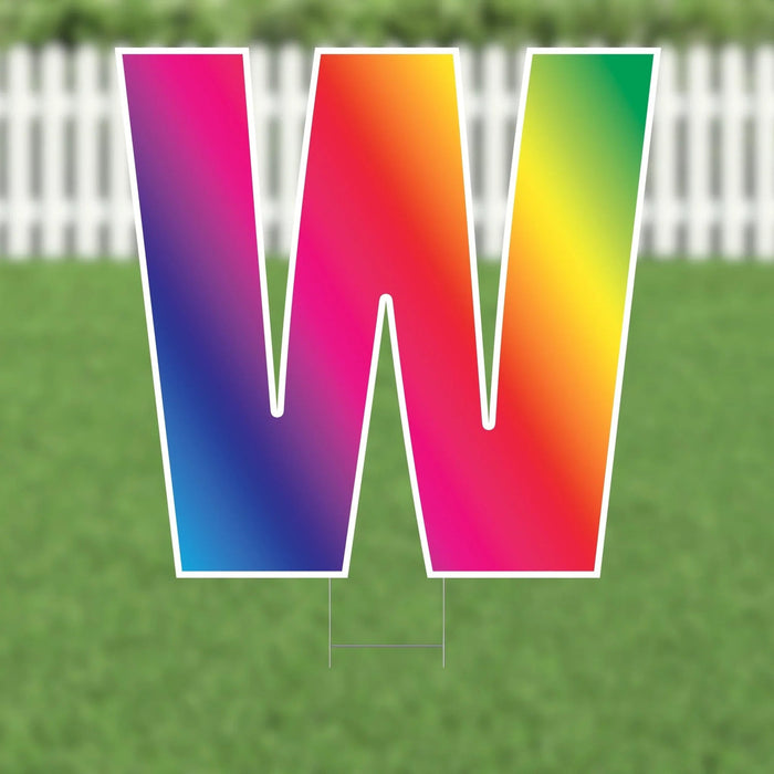 Colorful yard card letter W with gradient colors transitioning from purple and blue on the left to pink, red, orange, and yellow on the right, displayed on grass with a white picket fence background.