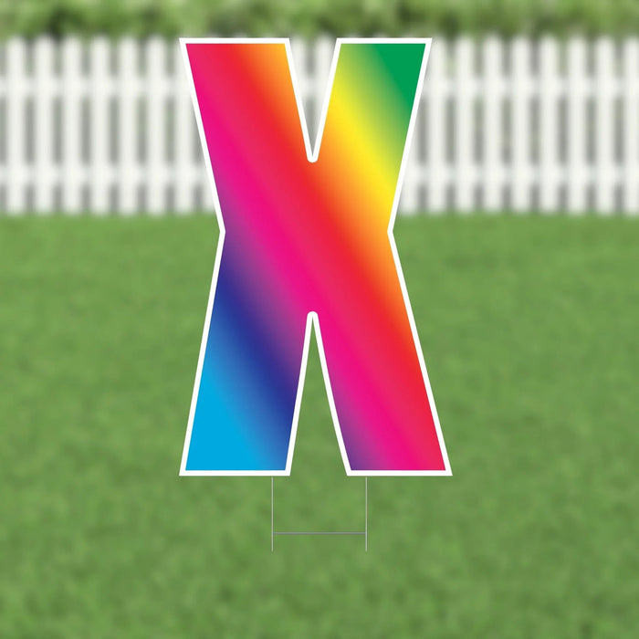 Colorful yard card letter X with a vibrant rainbow gradient transitioning from pink and orange at the top through yellow, green, blue, and purple, displayed on a lawn with white picket fence background.