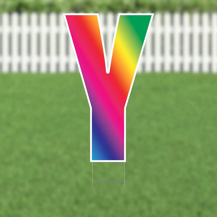 Colorful letter Y yard sign with rainbow gradient transitioning from pink and orange at the top through yellow, green, and blue to purple at the bottom, displayed on grass with a white picket fence background.