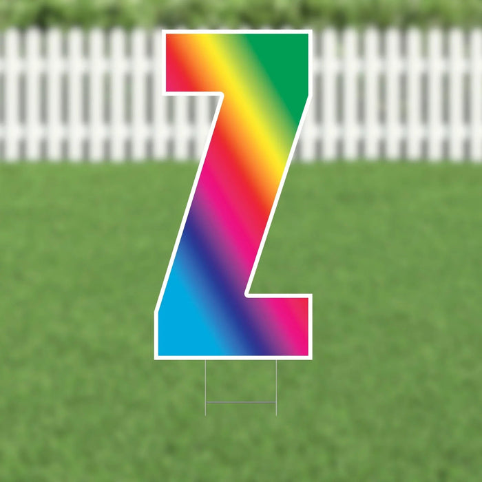 Rainbow gradient letter Z yard sign with white border and metal stakes, displayed on green grass with white picket fence background.