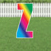 Rainbow gradient letter Z yard sign with white border and metal stakes, displayed on green grass with white picket fence background.
