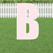 Pink letter B yard sign with white outline displayed on green grass with white picket fence background.