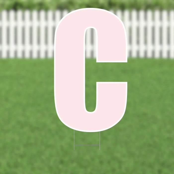 Large pink letter C yard sign with white outline displayed in grass with white picket fence in background.