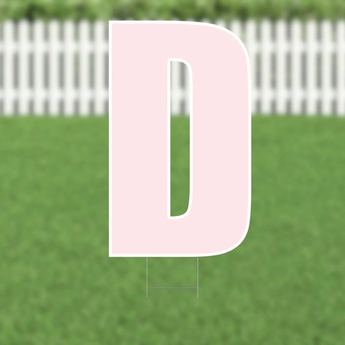 Pink letter D yard sign with white outline and metal stakes, displayed on grass with white picket fence background.