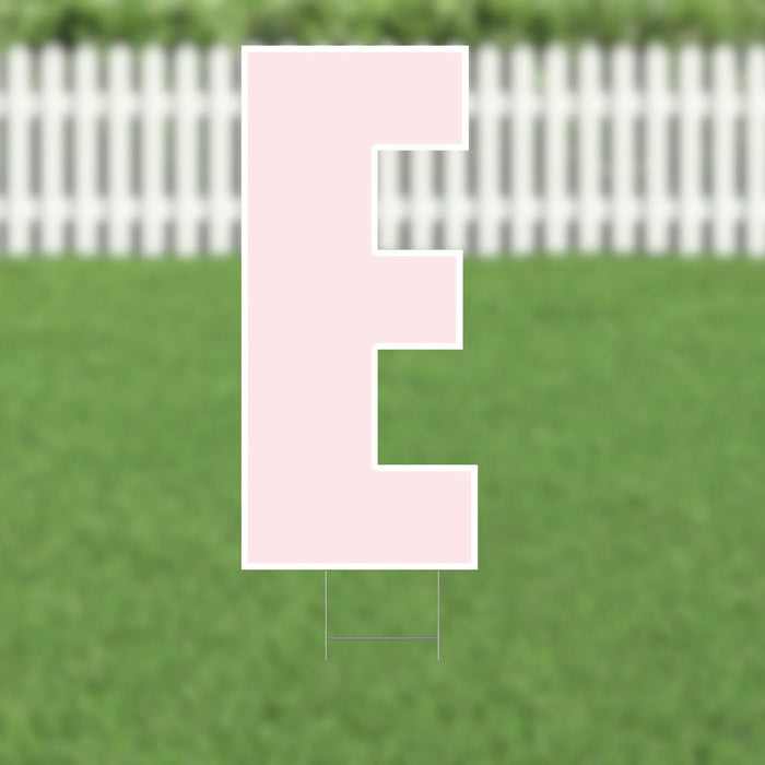 Pink letter E yard sign with white border and stake, displayed on green grass with white picket fence background.