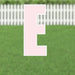 Pink letter E yard sign with white border and stake, displayed on green grass with white picket fence background.