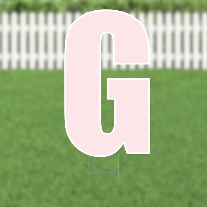 Large pink letter G yard sign with rounded edges displayed on a green lawn with white picket fence background.