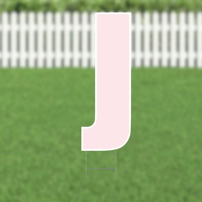 Pink letter J yard sign with white border and metal stakes, displayed on green grass with white picket fence backdrop.