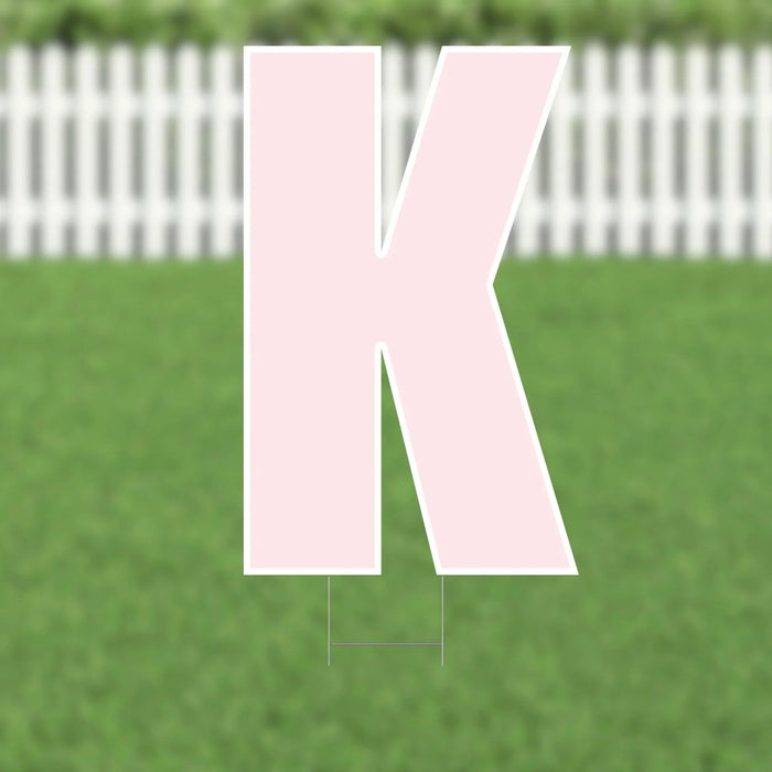 Large pink letter K yard sign with white border and supporting stakes, displayed on a green lawn with white picket fence in background.