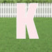 Large pink letter K yard sign with white border and supporting stakes, displayed on a green lawn with white picket fence in background.