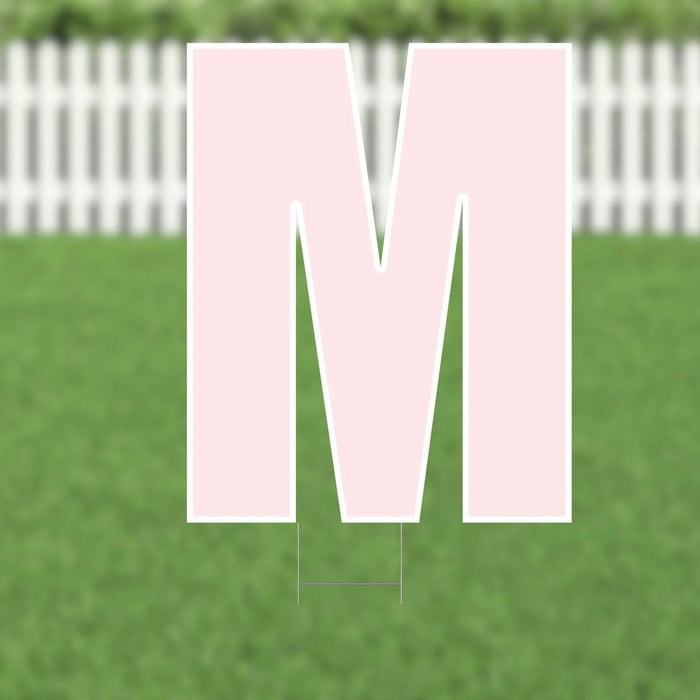 Pink corrugated plastic yard sign letter M, 21.5 inches tall, displayed on a residential lawn with white picket fence in background.