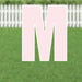 Pink corrugated plastic yard sign letter M, 21.5 inches tall, displayed on a residential lawn with white picket fence in background.