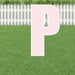 Large pink letter P yard sign with white border and metal stakes, displayed on a green lawn with white picket fence background.