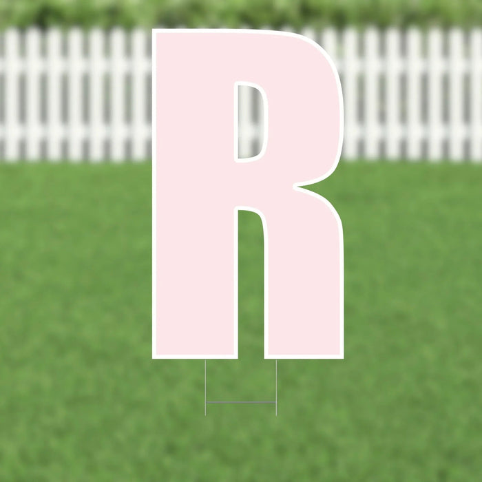 Large pink letter R yard sign with white outline and metal stakes, displayed on green grass with white picket fence background.