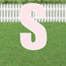 Pink letter S yard sign with white border and metal stakes, displayed on a green lawn with white picket fence in background.
