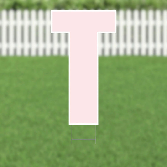 Pink letter T yard sign with white border and metal stakes, displayed on green grass with white picket fence in background.