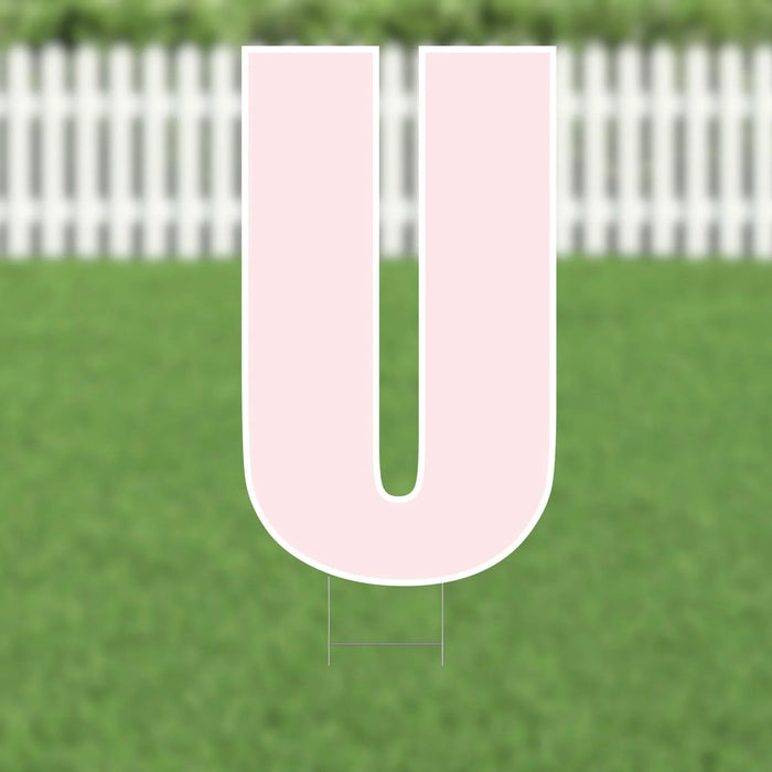Pink letter U yard sign with white border and metal stakes, displayed on green grass with white picket fence in background.