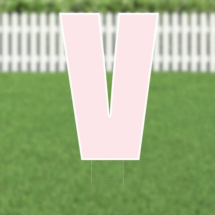 Large pink letter V yard sign with white border and metal stakes, displayed on green grass with white picket fence in background.