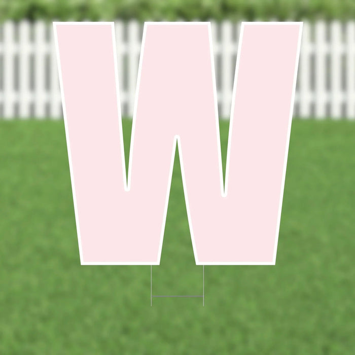 Large pink letter W yard sign with white border and metal stakes, displayed on grass with white picket fence in background.
