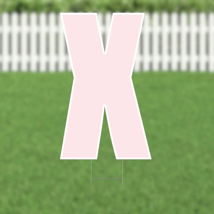 Large pink letter X yard sign with white border and metal stakes, displayed on green lawn with white picket fence in background.