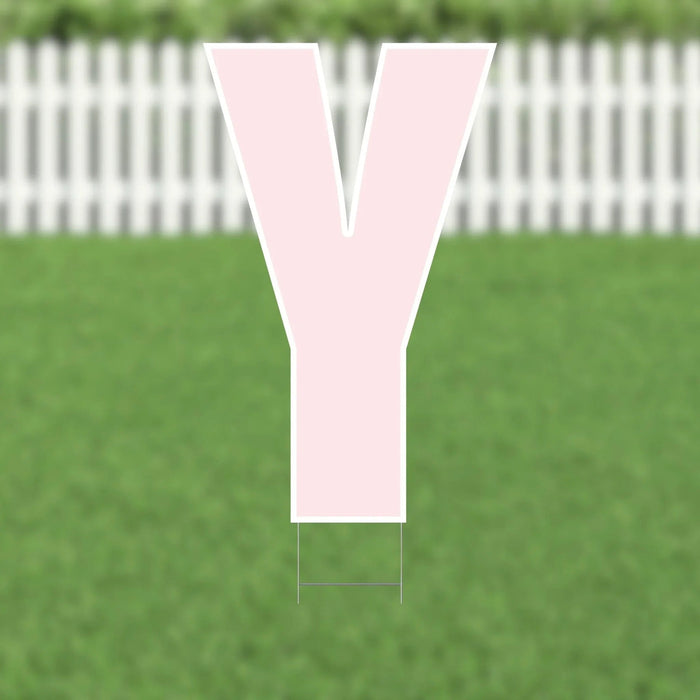 Large pink letter Y yard sign with white border and metal stakes, displayed on a lawn with white picket fence in background.
