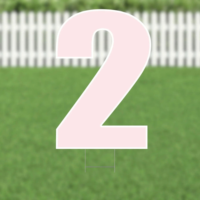 Large pink number 2 yard sign with metal stakes, displayed on green grass with a white picket fence in the background.