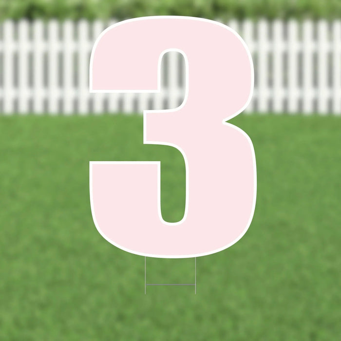 Large pink number 3 yard sign with white border and metal stakes, displayed on a lawn with white picket fence in background.