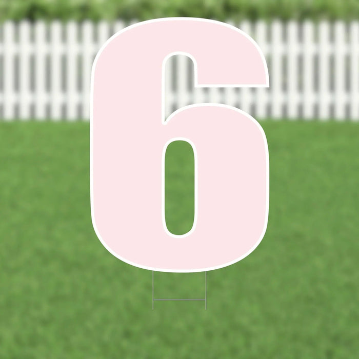 Large pink number 6 yard sign with white outline and metal stakes, displayed on green grass with white picket fence in background.