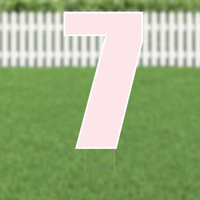 Pink number 7 yard sign with white outline and metal stakes, displayed on green grass with white picket fence background.