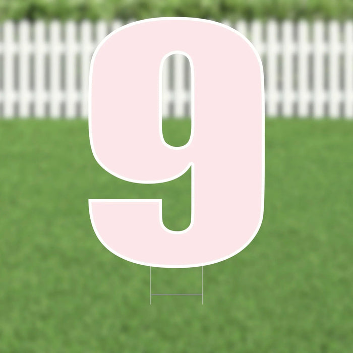 Large pink number 9 yard sign with white border and metal stakes, displayed on a green lawn with white picket fence in background.