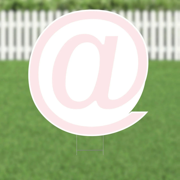 Large pink and white yard sign featuring an @ symbol, displayed on green grass with a white picket fence in the background.