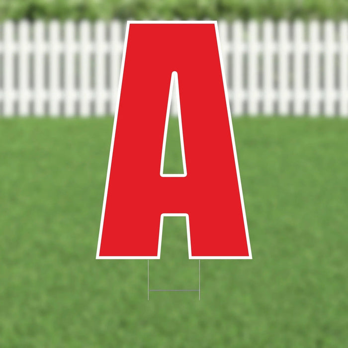 Large red letter A yard sign with white outline and cutout details, displayed on a grass lawn with white picket fence in background.