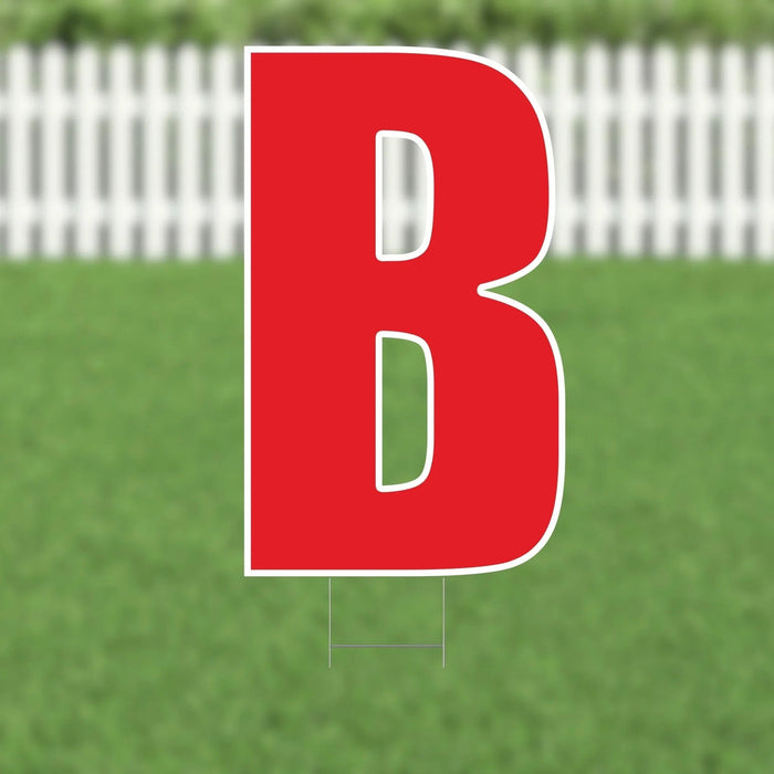 Large red letter B yard sign with white outline and metal stakes, displayed on green grass with white picket fence background.