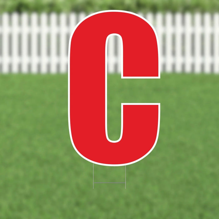 Large red letter C yard sign with white outline and metal stakes, displayed on green grass with white picket fence background.