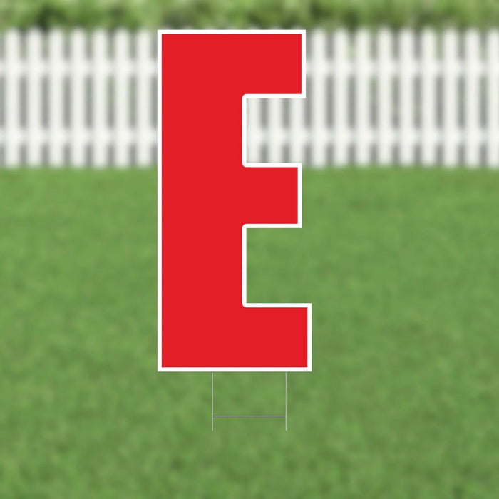 Red letter E yard sign with white outline and metal stakes, displayed on green grass with white picket fence background.