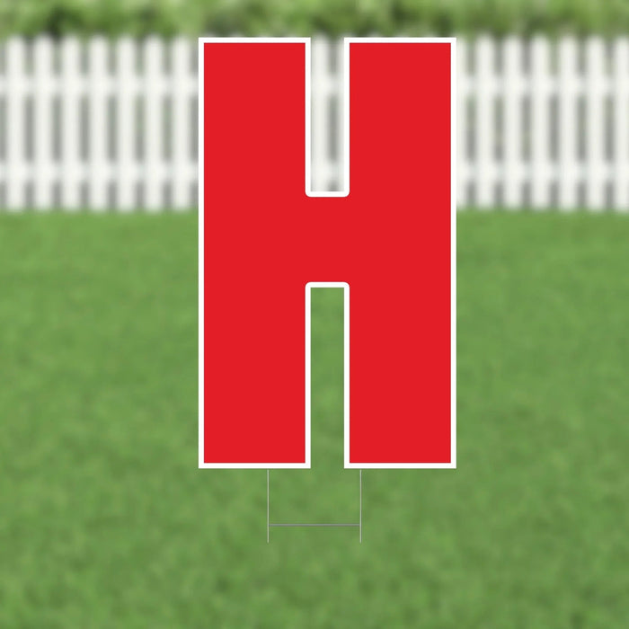 Red letter H yard sign with white border and metal stakes, displayed on a lawn with white picket fence in background.