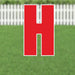 Red letter H yard sign with white border and metal stakes, displayed on a lawn with white picket fence in background.